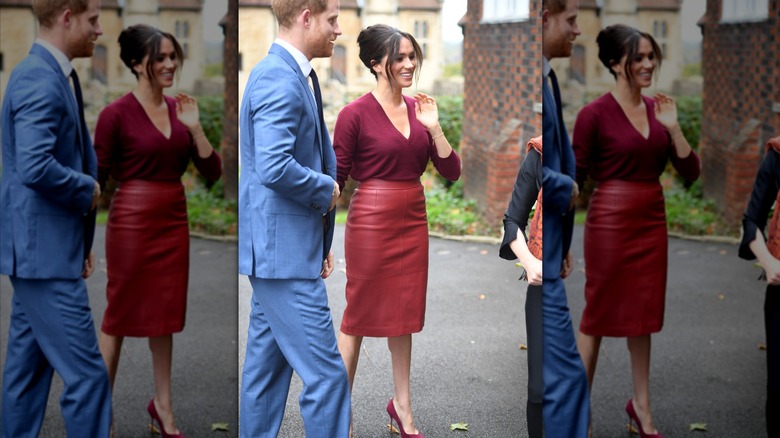Meghan Markle wearing a burgundy shirt and burgundy leather pencil skirt next Prince Harry in a blue suit.