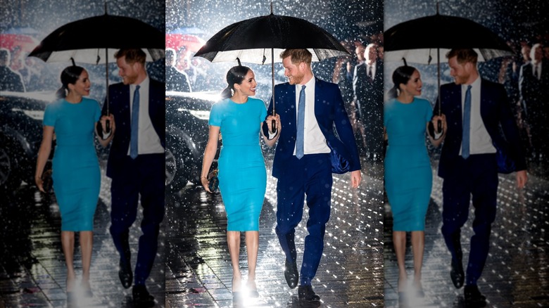 Meghan Markle and Prince Harry smiling at each other while holding an umbrella in the pouring rain, both wearing blue attire.