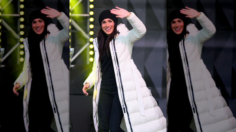 Meghan Markle wearing a black cap and a long white puffer jacket while waving and smiling.