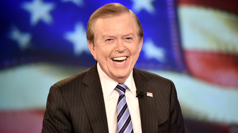 Lou Dobbs laughing on the set of Fox news against American flag backdrop