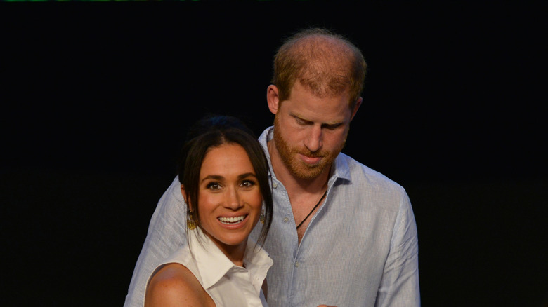 Meghan Markle smiling in front of Prince Harry in front of a black backdrop.