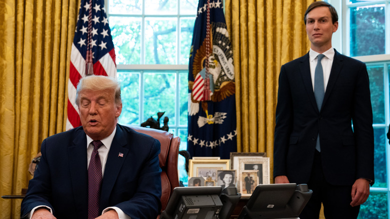 Jared Kushner standing behind a seated Donald Trump in the Oval Office.