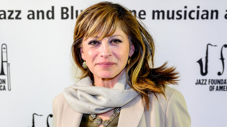 Maria Bartiromo in a coat and scarf standing in front of a white backdrop.