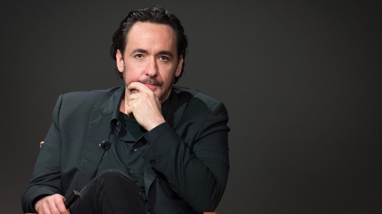 John Cusack resting his chin in his hands while onstage in the mid-2010s