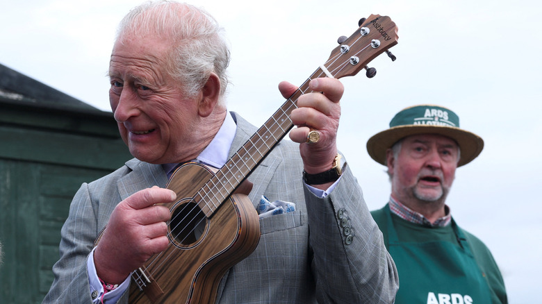 King Charles III playing a ukulele while another man looks on behind him