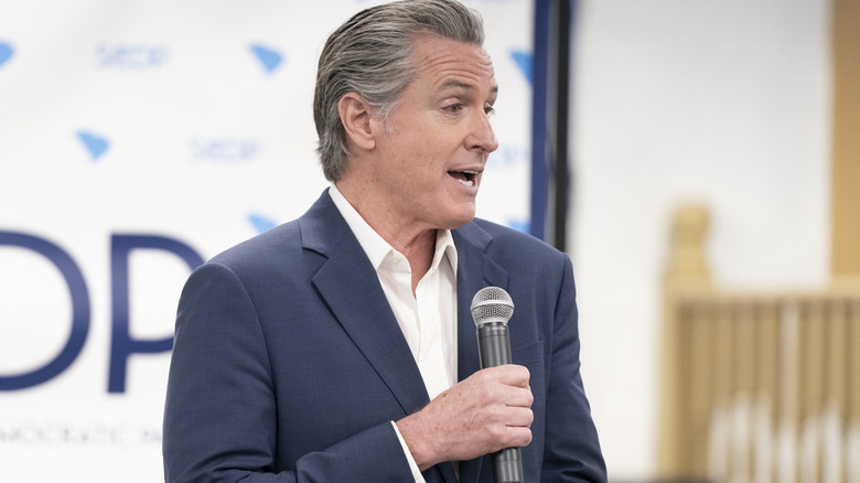 Gavin Newsom delivering a speech in a grey suit with an open-collared white shirt