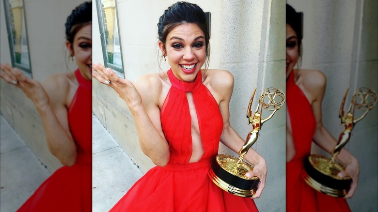 A shocked smiling Kate Mansi holds a gold Emmy Award in a red dress