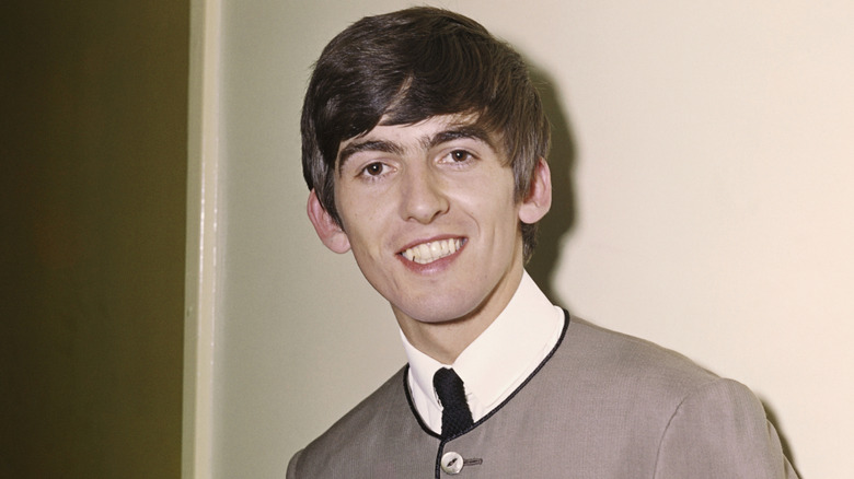 George Harrison smiling while posing for a photo in a beige blazer, white shirt, and a black tie