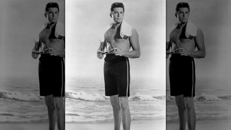 Max Baer Jr. in dark swim trunks posing before a beach