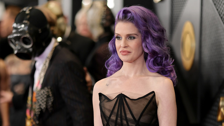 image of Kelly Osbourne with a weird expression on the red carpet of an event while Sid Wilson with a black mask stands away from her and out of focus