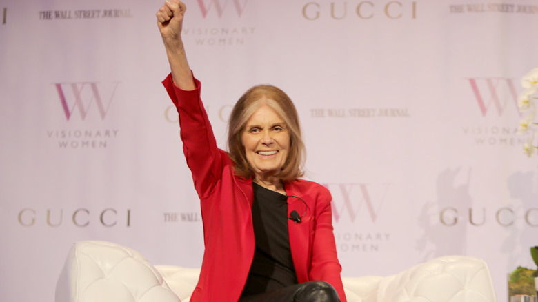 Gloria Steinem lifting her fist into the air in a red blazer onstage