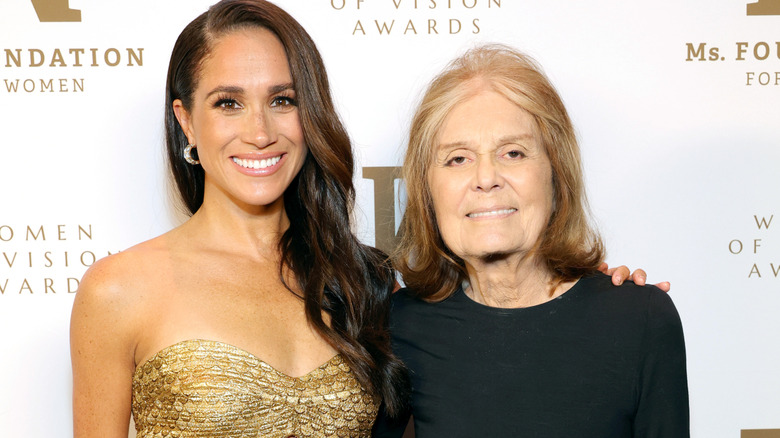 A smiling Meghan Markle and Gloria Steinem smiling while posing standing arm in arm