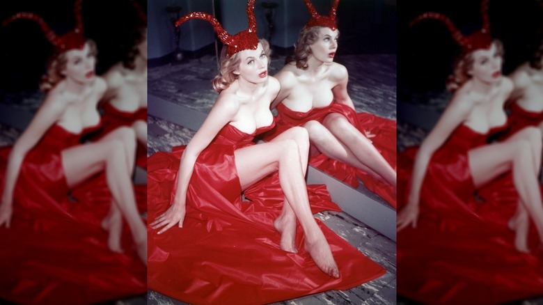 Swedish actress Anita Ekberg sitting by a mirror in a red satin dress and a sequinned devil-horn cap, circa 1956.
