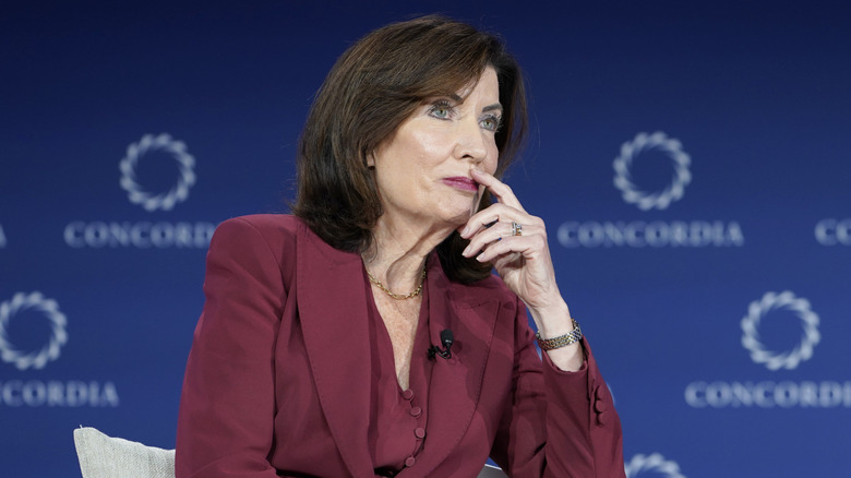 Kathy Hochul in a maroon suit, sitting in a white chair with her hand to her face.