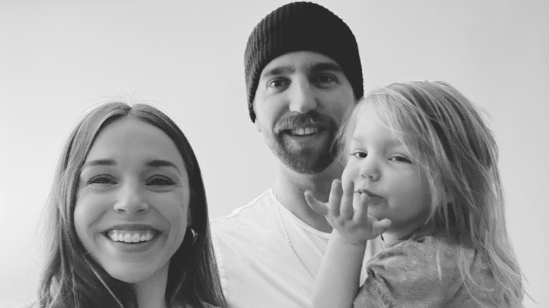 Actor  Elise Gatien smiles in a selfie next to her fiancé who wears a beanie and holds their young child