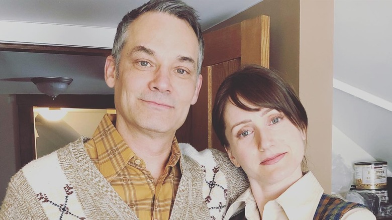 Meghan Gardiner stands inside in front of a door half smiling next to her husband Todd Thomson and both wear collared shirts