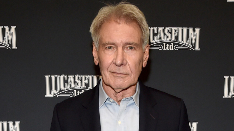 Harrison Ford posing in front of Lucasfilm backdrop in a black suit jacket and a light-blue open-collar shirt