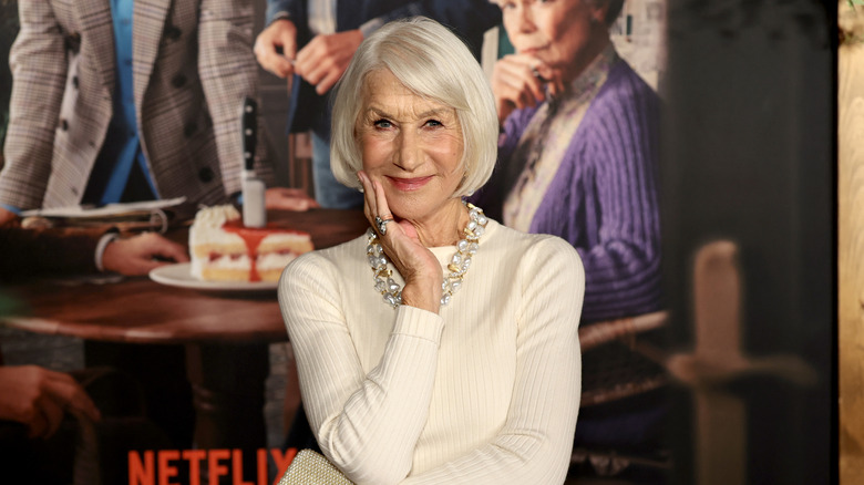 Helen Mirren with her hand gentle on her face, smiling while wearing an all-white outfit.