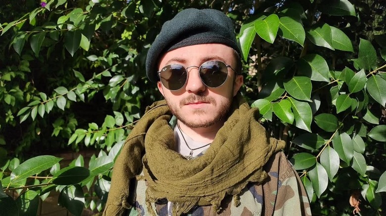 Close-up of Atticus Shaffer wearing a hat, sunglasses, and a camouflage jacket and standing in front of a bush
