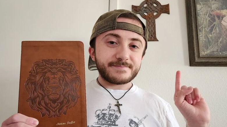 Atticus Shaffer holding a journal with a lion and his name on the cover and pointing to a cross on the wall behind him