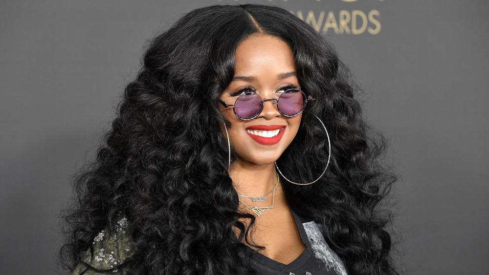 H.E.R. smiling at event, wearing purple-tinted glasses