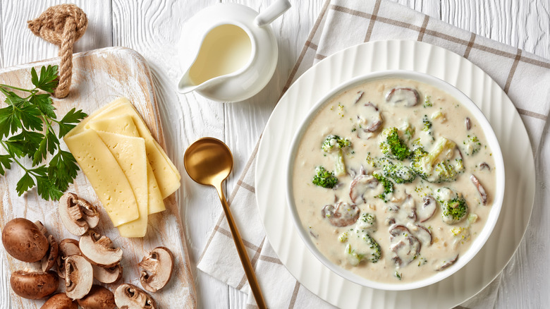 Mushroom Broccoli Cheese Soup in a white bowl with a golden spoon and ingredients on a white wooden table, horizontal view from above, close-up