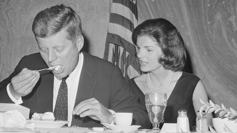 Jackie Kennedy staring at JFK eating a piece of dessert at a formal lunch