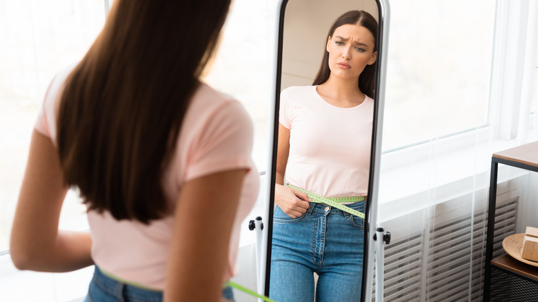 Woman measures her waist with a tape measurer, looks sad in mirror