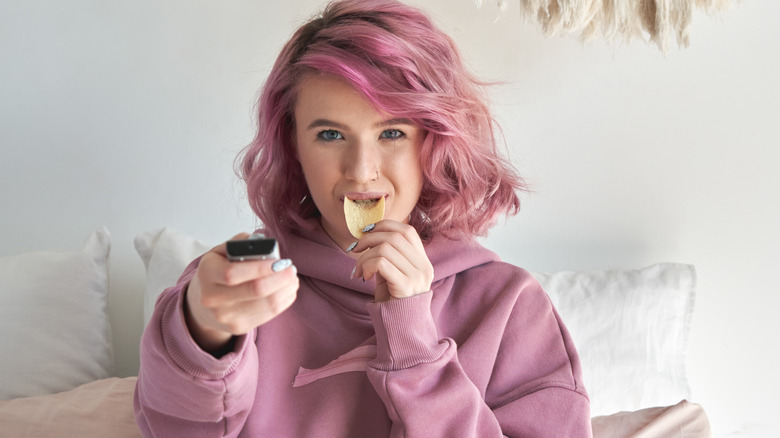 A woman eating chips in bed with a remote in her hand 