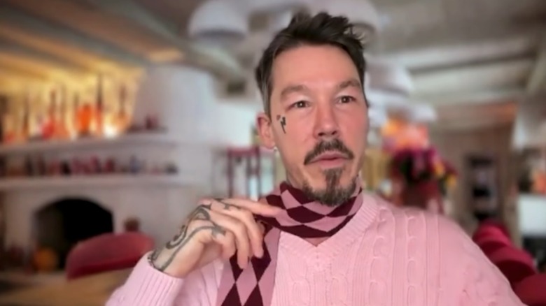David Bromstad wearing a pink sweater and red and pink scarf sitting in his living room