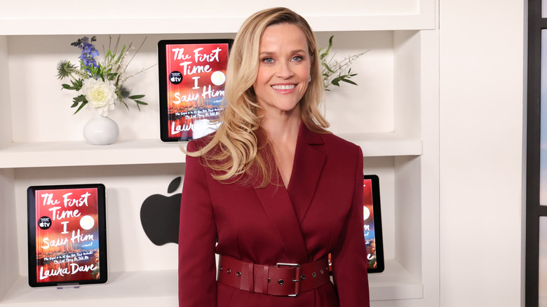 Reese Witherspoon smiling and wearing a reddish maroon outfit in front of a digital book display