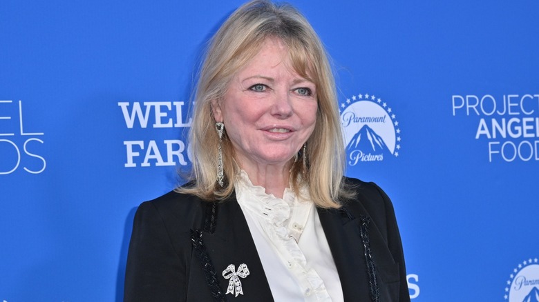 Cheryl Tiegs posing wearing a white shirt and black suit jacket on a red capet.