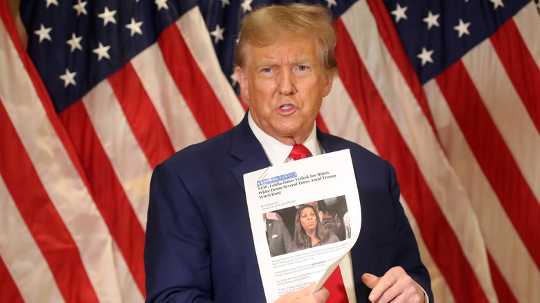 Donald Trump holding a printout with Letitia James' face on it in front of American flags