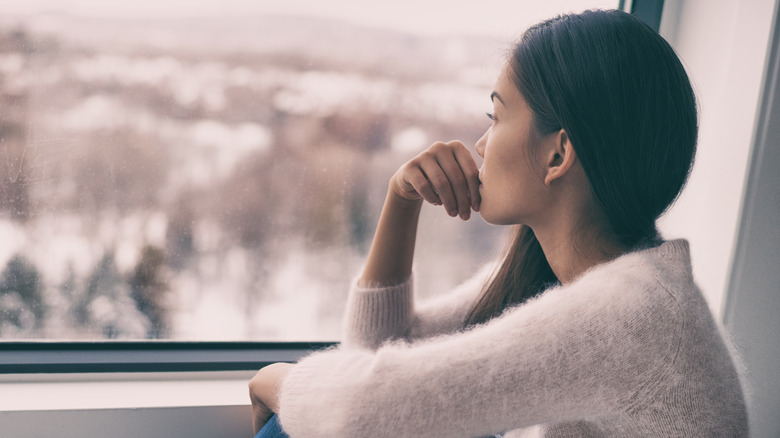 Sad pensive woman at home seasonal depression - seasonal affective disorder mental health looking out the window alone.