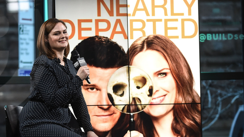 Emily Deschanel sits holding a microphone in a black polka dot dress in front of a screen with her character in "Bones" on it