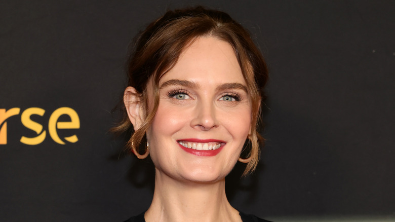 Emily  Deschanel smiles with her hair pulled back and tendrils hanging down wearing gold hoops on the red carpet