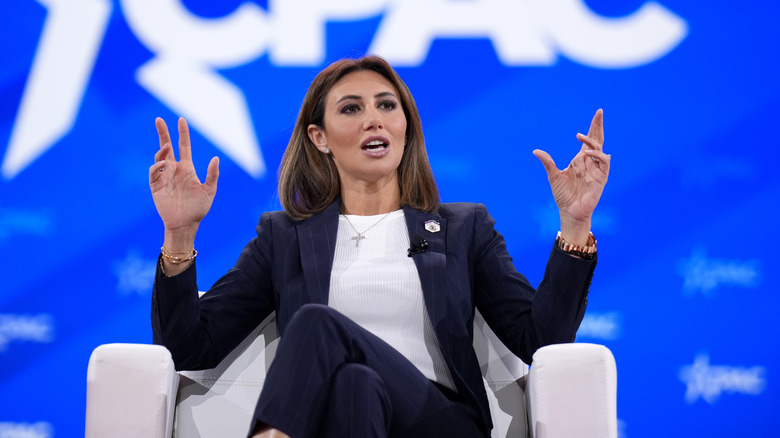 Alina Habba raising her hands while speaking onstage in a black blazer with a white top underneath