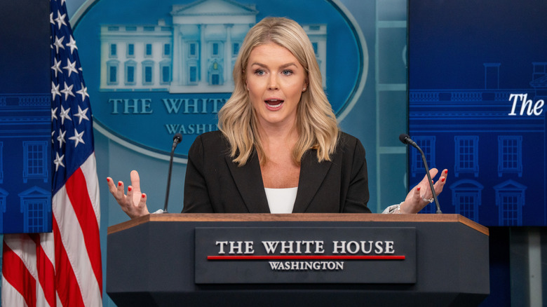 Karoline Leavitt raising her hands while speaking behind a podium in a black blazer and a white shirt