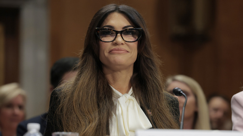 Kimberly Guilfoyle smiling at a hearing while wearing glasses and a white blouse