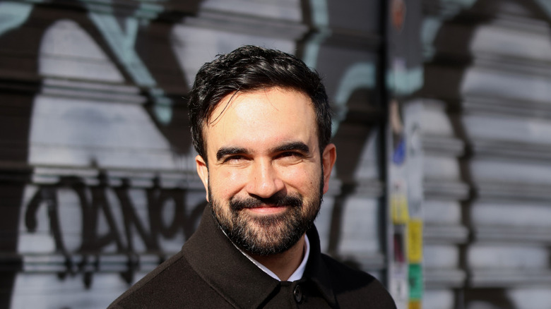 Zohran Mamdani in a black jacket, smiling in front of a graffiti riddled wall.