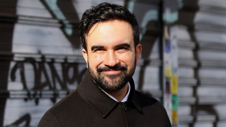 Zohran Mamdani in a black jacket, smiling in front of a graffiti riddled wall.