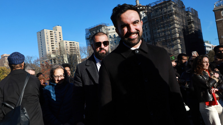 Crooked angle of Zohran Mamdani in a black coat, surrounded by people.