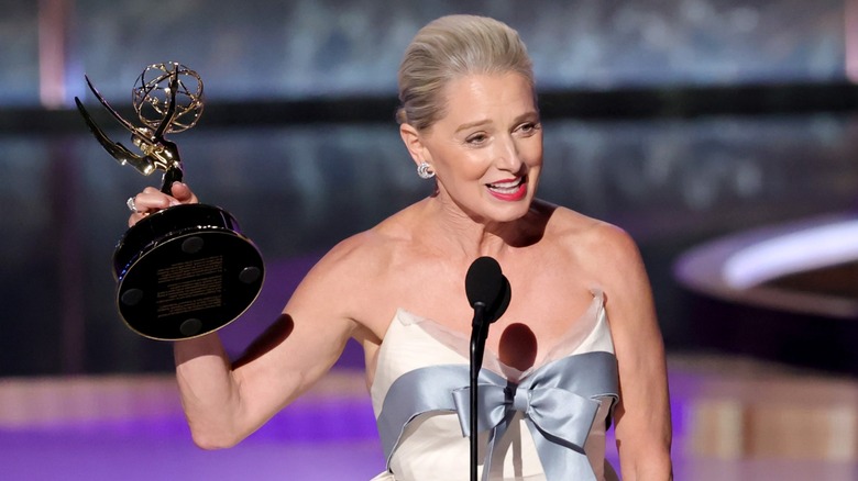 Katherine LaNasa in a cream dress with a silver bow on it, holding up an Emmy award as she speaks into a mic.