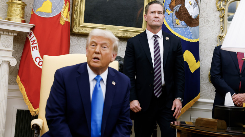 Donald Trump sitting in the Oval Office, slightly out of focus, with Mike Waltz standing behind him in focus