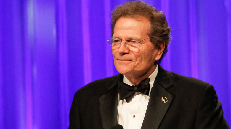 Patrick Wayne wearing a suit and standing in front of a purple background