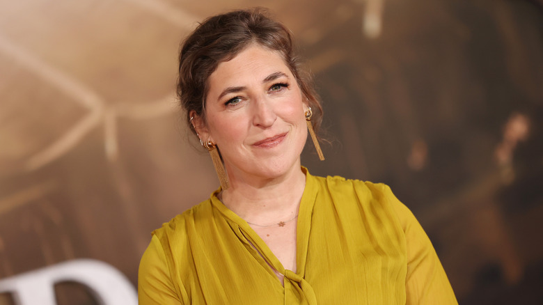Mayim Bialik at a red carpet event wearing mustard-yellow dress and drop earrings.