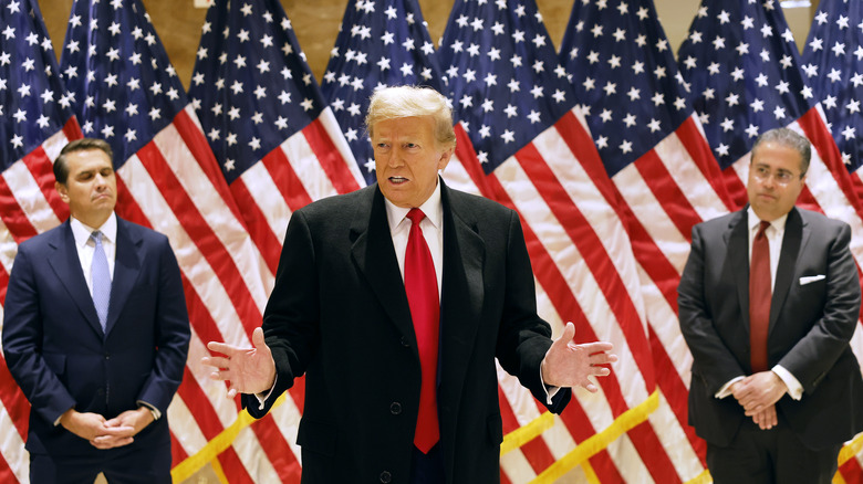 Trump talking with arms spread American flags in background