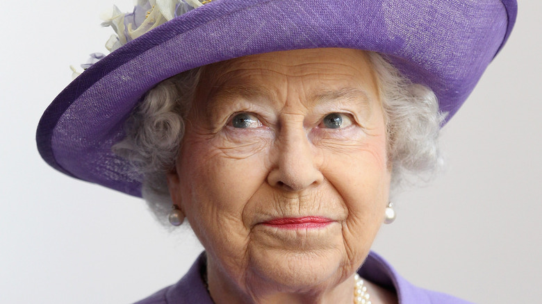 Queen Elizabeth II smiling and looking sideways in a lavender hat and pearls