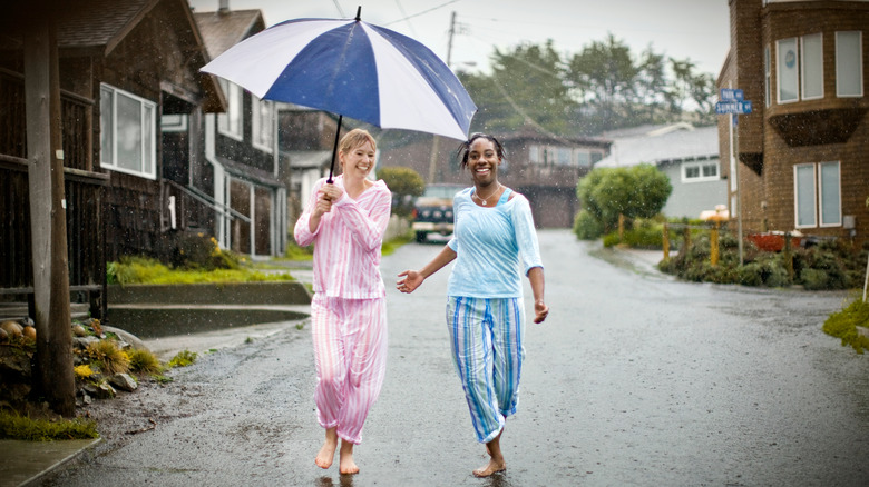 Two women wearing pajamas while walking down the street holding an unbrella.