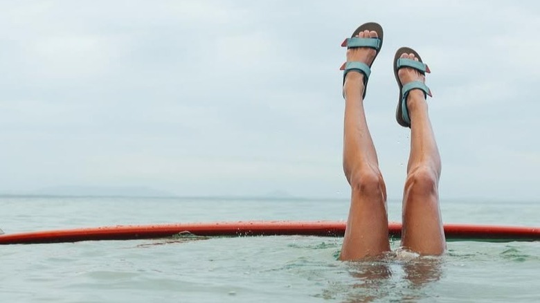 A pair of legs sticking out of the ocean, with the feet wearing teal sandals